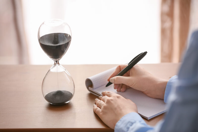 Woman writing something in notebook on table with hourglass in office. Time management concept