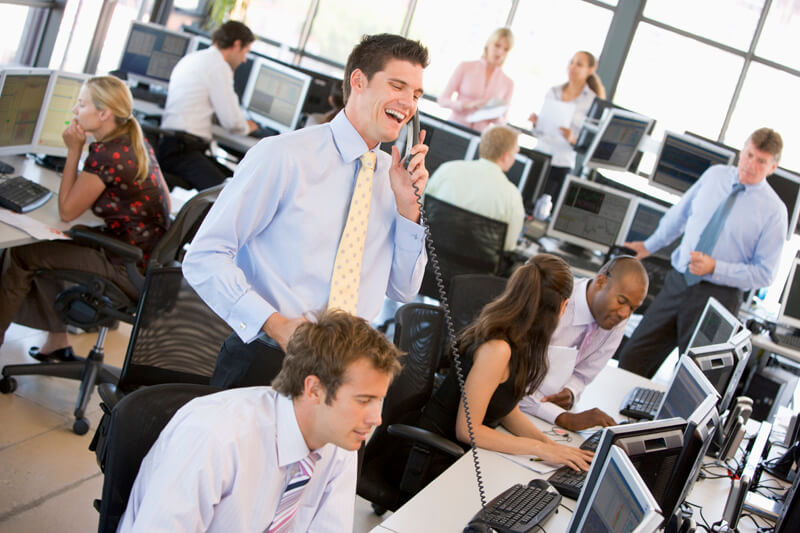 View Of Busy Office with most employees sitting, one man standing talking on phone