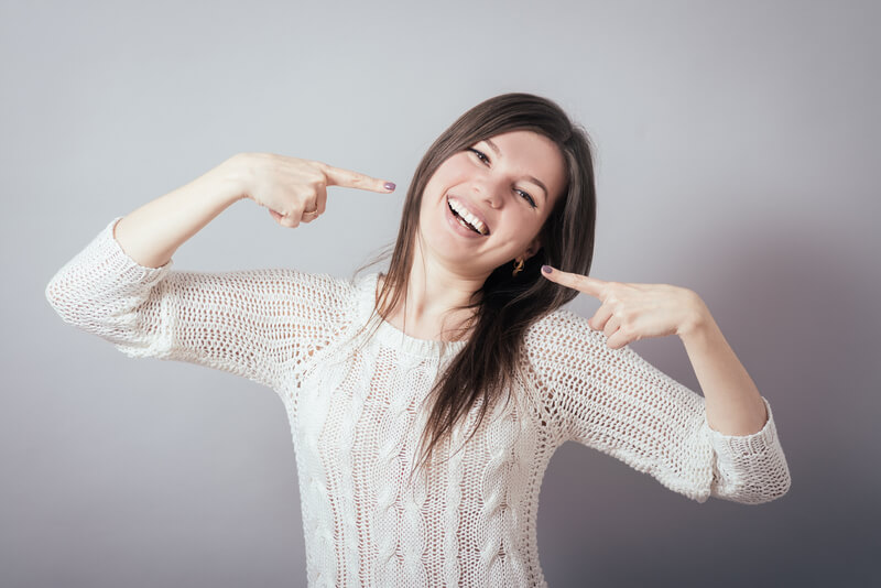 woman in white sweater pointing at her face with both hands and laughing