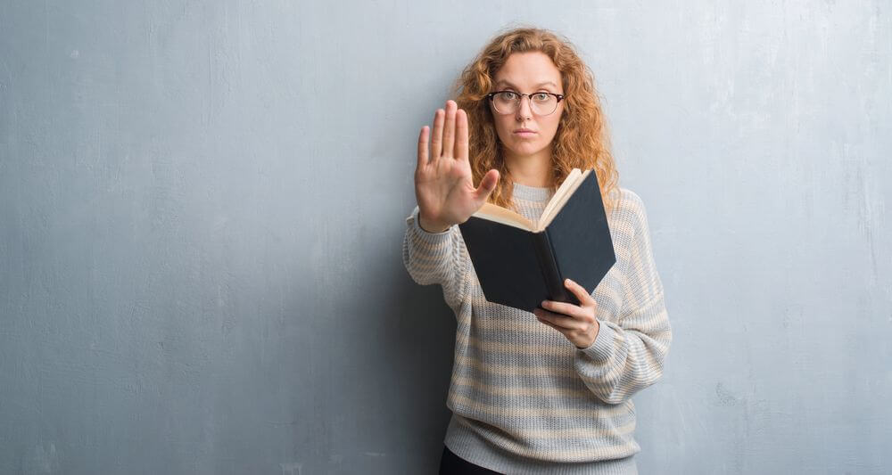 Young redhead woman over grey grunge wall reading a book with open hand doing stop sign with serious and confident expression, defense gesture